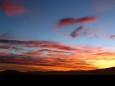 The moon appears in a blue sky amongst yellow, orange, and red clouds lit during a sunset.